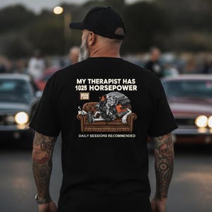 May include: A black t-shirt featuring a graphic on the back of a car engine sitting on a brown couch, holding a coffee mug. The text reads: MY THERAPIST HAS 1025 HORSEPOWER and DAILY SESSIONS RECOMMENDED. A small framed certificate is visible above the couch.