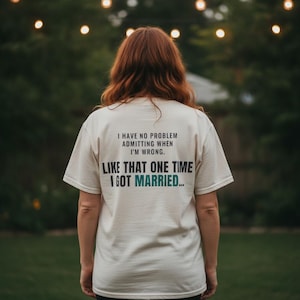 May include: A cream-colored short-sleeve t-shirt featuring black and teal text on the back that reads: I HAVE NO PROBLEM ADMITTING WHEN I'M WRONG. LIKE THAT ONE TIME I GOT MARRIED...