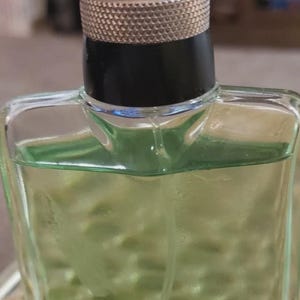 May include: A clear glass perfume bottle with a silver cap and black collar. The bottle contains a light green liquid. The cap has a textured, ridged design. The bottle is square with rounded edges.
