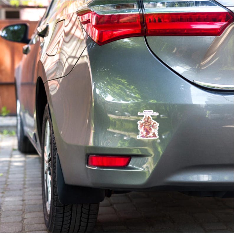 May include: A rectangular sticker applied to the rear bumper of a gray car. The sticker features a colorful illustration of the Hindu deity Ganesha surrounded by floral elements, with the text "GANESHA" printed in black above the deity figure.