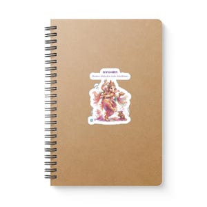 May include: A spiral-bound notebook with a plain tan cover featuring a central sticker. The sticker displays a colorful illustration of Ganesha with the text "GANESHA" and "Remove obstacles, invite abundance." A small mouse is depicted near Ganesha's feet.