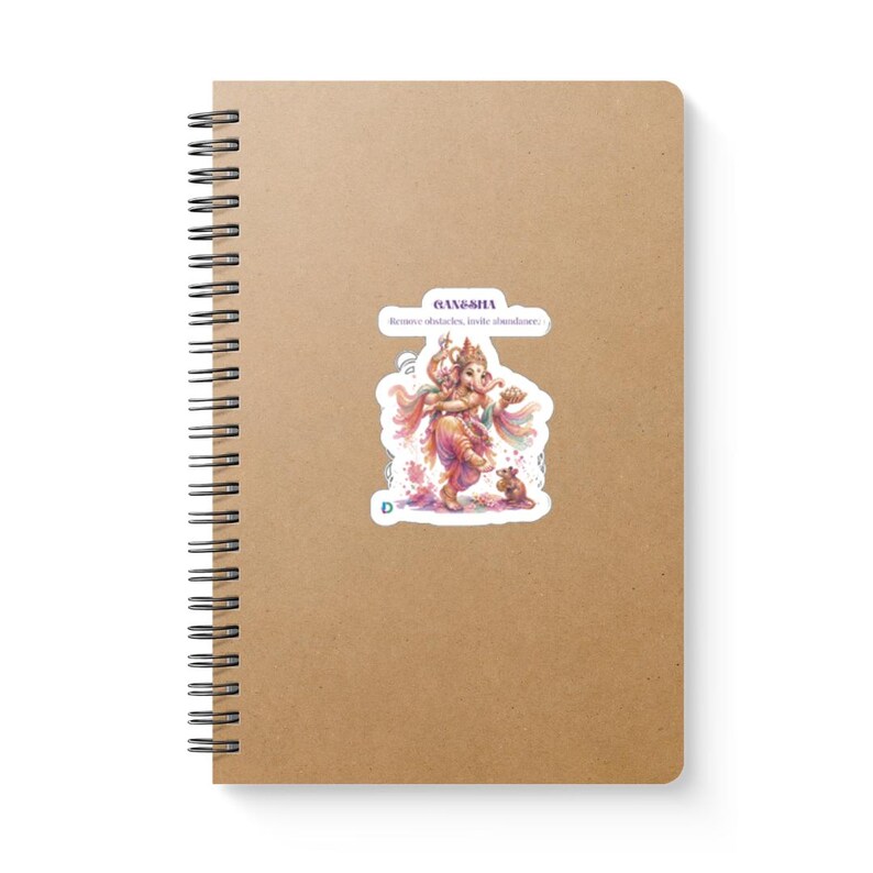 May include: A spiral-bound notebook with a plain tan cover featuring a central sticker. The sticker displays a colorful illustration of Ganesha with the text "GANESHA" and "Remove obstacles, invite abundance." A small mouse is depicted near Ganesha's feet.