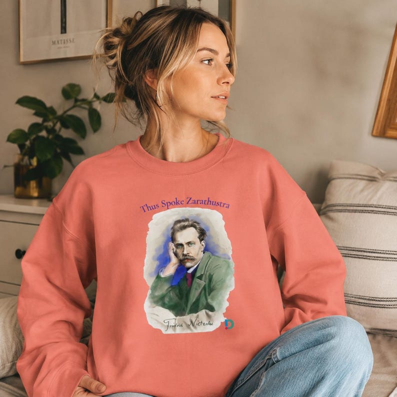 Peut inclure: Un sweat-shirt &agrave; col rond de couleur corail avec une impression graphique de Friedrich Nietzsche portant une veste verte. Le texte "Thus Spoke Zarathustra" est imprim&eacute; en violet au-dessus du portrait, avec "Friedrich Nietzsche" &eacute;crit en dessous.