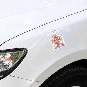 May include: A white car fender features a colorful sticker depicting the Hindu deity Ganesha in a dancing pose, adorned in orange and pink garments with flowing scarves, accompanied by a small mouse, with text printed above the figure.