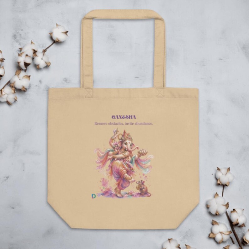 Op de afbeelding: Een beige draagtas met een kleurrijke illustratie van de godheid Ganesha die danst met een kleine muis. De tekst Ganesha en Remove obstacles, invite abundance staat erboven gedrukt, met een kleine groene letter D in de hoek.