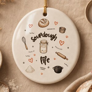 Può includere: Un ornamento in ceramica color crema di forma rotonda appeso a un cordino dorato. La superficie presenta illustrazioni marroni di utensili da cucina, una pagnotta di lievito madre, un barattolo e una pentola, con il testo "Sourdough Life".