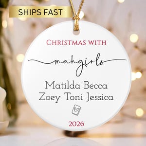Puede incluir: Un adorno redondo de cerámica blanca colgado de un cordón dorado. El texto dice CHRISTMAS WITH mahgirls, seguido de los nombres Matilda Becca, Zoey Toni Jessica, un pequeño icono de corazón en un cuadrado y el año 2026 en color rojo.