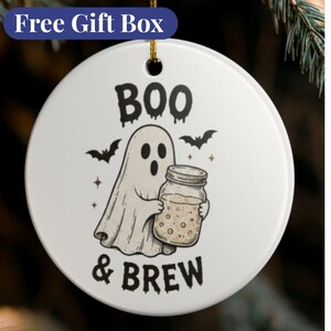 Può includere: Un ornamento rotondo in ceramica bianca con una grafica nera di un fantasma che tiene in mano un barattolo di vetro pieno di un liquido beige. Il testo "BOO & BREW" è stampato in nero sopra e sotto il fantasma, con piccole illustrazioni di pipistrelli.