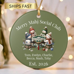 May include: A round olive green ceramic ornament featuring a gold hanging cord. The centre displays four snowmen playing mahjong at a table. Text reads: Merry Mahj Social Club, Tom, Evelyn, Charlie, Becca, Nash, Toby, and Est. 2026 in white font.