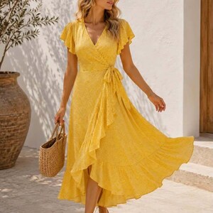 Peut inclure: Une robe portefeuille jaune à motifs floraux et bordure à volants. La robe a des manches courtes à volants et une ceinture à la taille. Un sac fourre-tout en paille et des sandales complètent le look. Le texte "Amara Riviera" est visible.