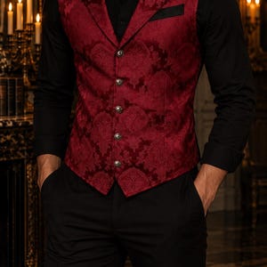 Può includere: Un gilet rosso scuro con un motivo damascato tono su tono, revers dentellato e cinque bottoni metallici sul davanti. Il gilet è indossato sopra una camicia nera a maniche lunghe e abbinato a pantaloni neri, creando un completo formale.