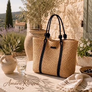 May include: A woven straw tote bag with black rope handles. The bag has a rectangular shape and a small black tag. The text "Amara Riviera" is at the bottom of the image. The bag is on a stone surface.