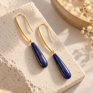 Peut inclure: Une paire de boucles d'oreilles pendantes composées de pierres de lapis-lazuli allongées en forme de goutte, d'un bleu profond avec des éclats dorés. Les pierres sont suspendues à des crochets en métal doré poli aux lignes courbes et épurées.