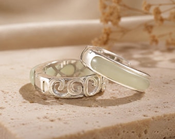 May include: Two silver rings featuring a smooth, pale green stone inlay. One ring displays a textured, swirling pattern along the band, while the other has a polished, minimalist silver finish. Both rings are crafted with a metallic silver band.