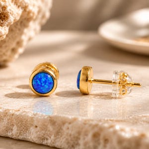 May include: A pair of stud earrings featuring round, vibrant blue opal cabochons set in polished gold-tone metal bezels. Each earring includes a thin gold-tone post and a clear, circular plastic backing for secure attachment to the earlobe.