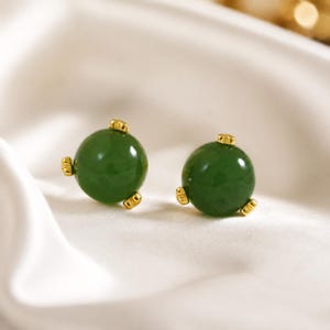 May include: A pair of stud earrings featuring smooth, spherical dark green jade stones. Each stone is secured by three small, polished gold-tone metal prongs that grip the surface of the sphere, creating a simple and clean design for these jewelry pieces.