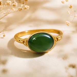 Puede incluir: Un anillo de oro con una piedra preciosa de color verde oscuro, lisa y de forma ovalada, engastada en el centro. La banda de metal tiene un acabado pulido con detalles decorativos en los lados donde la banda se une al engaste central.