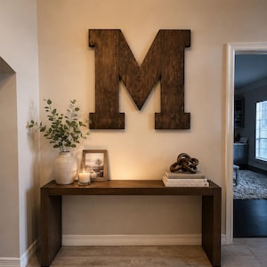 May include: A large, dark brown wooden letter M mounted on a wall above a rectangular dark wood console table. The table holds a ceramic vase with green stems, a framed picture, a glass candle, a stack of two books, and a brown decorative knot.