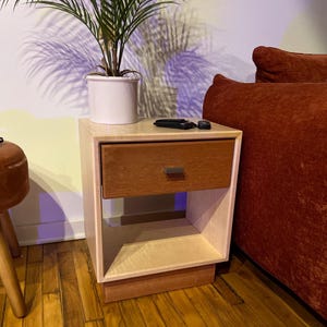 May include: A two-toned wooden nightstand with a drawer and open shelf. The drawer is a warm brown, while the frame is a lighter wood. A small plant in a white pot sits on top, along with a remote control.