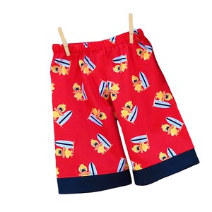 May include: Red shorts with a pattern of yellow ducks riding surfboards. The shorts have a navy blue trim.