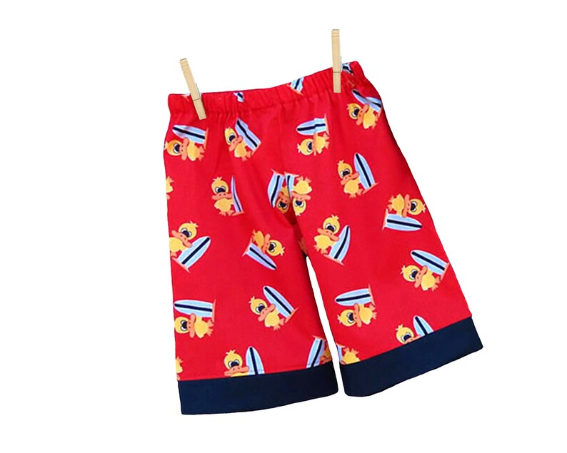 May include: Red shorts with a pattern of yellow ducks riding surfboards. The shorts have a navy blue trim.
