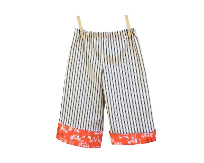 May include: White and black striped pants with orange trim featuring a bicycle print. The pants are hanging on a clothesline with wooden clothespins.