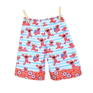 May include: Blue and white striped shorts with a red Elmo print. The shorts have a red and orange trim with a circle pattern.
