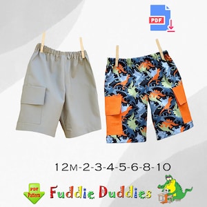 Puede incluir: Dos pares de pantalones cortos para niños, un par es gris y el otro es azul con estampado de dinosaurios. Los pantalones cortos tienen bolsillos y están disponibles en las tallas 12M-2-3-4-5-6-8-10. El texto "Fuddie Duddies" está en la imagen.