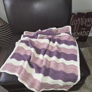 Pode incluir: Uma manta feita &agrave; m&atilde;o em croch&ecirc; com um padr&atilde;o ondulado repetitivo em listras horizontais alternadas de fio rosa seco, roxo ameixa profundo e branco off-white. As bordas do tecido retangular s&atilde;o finalizadas com uma borda simples.