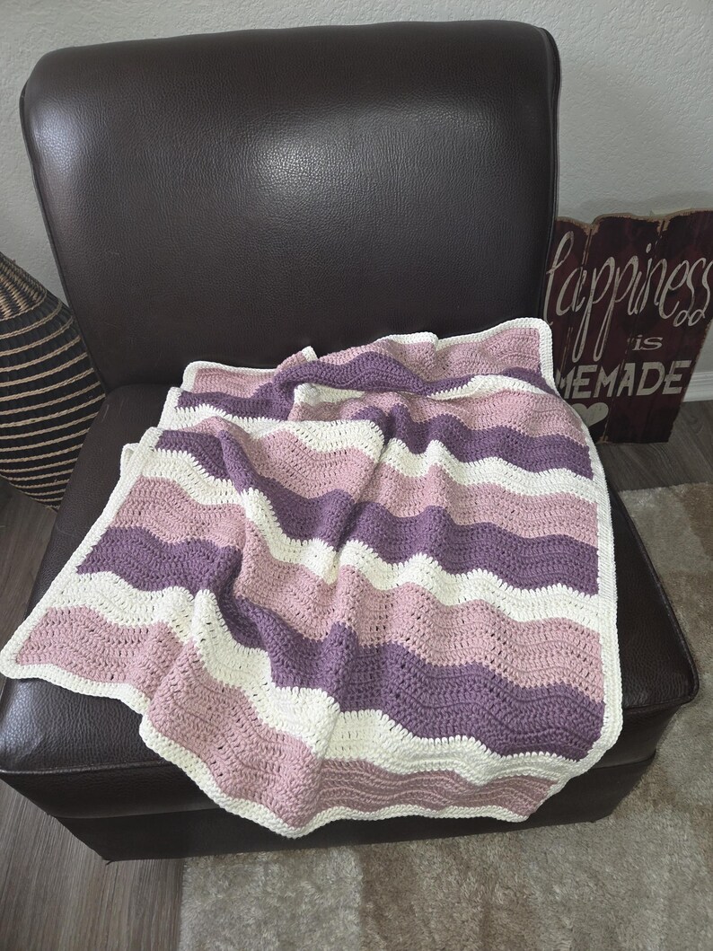 Pode incluir: Uma manta feita &agrave; m&atilde;o em croch&ecirc; com um padr&atilde;o ondulado repetitivo em listras horizontais alternadas de fio rosa seco, roxo ameixa profundo e branco off-white. As bordas do tecido retangular s&atilde;o finalizadas com uma borda simples.