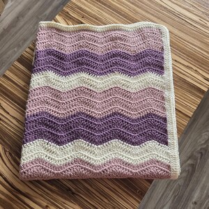 Pode incluir: Uma manta retangular de croch&ecirc; com um padr&atilde;o repetitivo de ondas em zigue-zague em listras horizontais alternadas de creme, rosa antigo e roxo escuro. A manta &eacute; finalizada com uma borda de cor creme s&oacute;lida em todos os quatro lados.