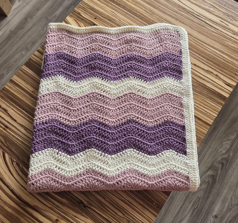 Pode incluir: Uma manta retangular de croch&ecirc; com um padr&atilde;o repetitivo de ondas em zigue-zague em listras horizontais alternadas de creme, rosa antigo e roxo escuro. A manta &eacute; finalizada com uma borda de cor creme s&oacute;lida em todos os quatro lados.