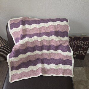 Pode incluir: Uma manta de croch&ecirc; retangular com um padr&atilde;o repetitivo de ondas ou chevron. O design consiste em listras horizontais em cores alternadas creme, rosa antigo e malva profundo, criando uma apar&ecirc;ncia suave, texturizada e em camadas.