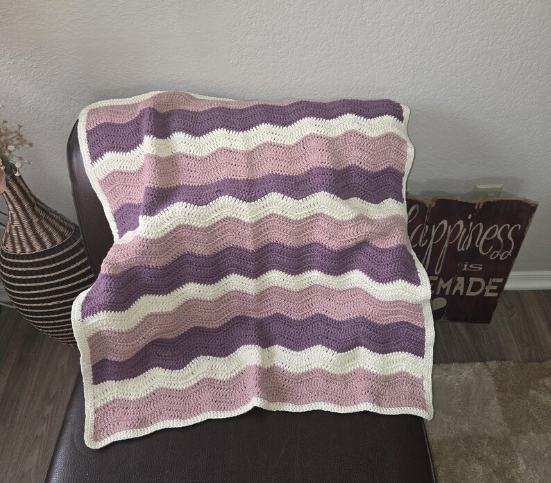 Pode incluir: Uma manta de croch&ecirc; retangular com um padr&atilde;o repetitivo de ondas ou chevron. O design consiste em listras horizontais em cores alternadas creme, rosa antigo e malva profundo, criando uma apar&ecirc;ncia suave, texturizada e em camadas.
