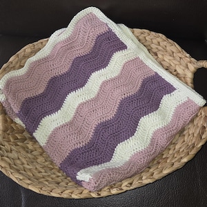 Pode incluir: Uma manta de croch&ecirc; dobrada com um padr&atilde;o ondulado repetitivo em listras alternadas de creme, rosa antigo e ameixa escuro. O tecido &eacute; feito com um ponto consistente, apresentando uma textura macia e uma borda limpa e bem acabada.