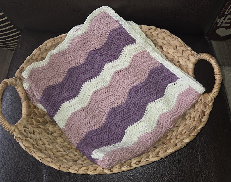 Pode incluir: Uma manta de croch&ecirc; dobrada com um padr&atilde;o ondulado repetitivo em listras alternadas de creme, rosa antigo e ameixa escuro. O tecido &eacute; feito com um ponto consistente, apresentando uma textura macia e uma borda limpa e bem acabada.
