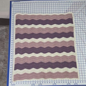 Pode incluir: Uma manta de croch&ecirc; retangular com um padr&atilde;o ondulado em listras alternadas de creme, rosa antigo e roxo escuro. A manta est&aacute; posicionada sobre uma base de corte, medindo aproximadamente 66 cm de largura e 76 cm de comprimento.