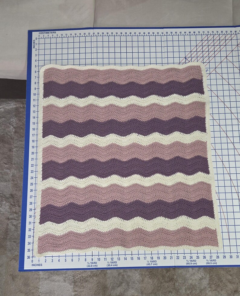Pode incluir: Uma manta de croch&ecirc; retangular com um padr&atilde;o ondulado em listras alternadas de creme, rosa antigo e roxo escuro. A manta est&aacute; posicionada sobre uma base de corte, medindo aproximadamente 66 cm de largura e 76 cm de comprimento.