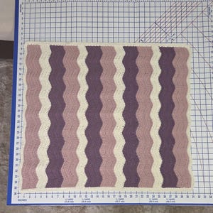 Pode incluir: Uma manta retangular de croch&ecirc; com um padr&atilde;o ondulado e listras verticais alternadas em creme, rosa antigo e roxo escuro. A manta est&aacute; posicionada sobre uma base de corte com medidas m&eacute;tricas, medindo aproximadamente 61 cm por 66 cm.