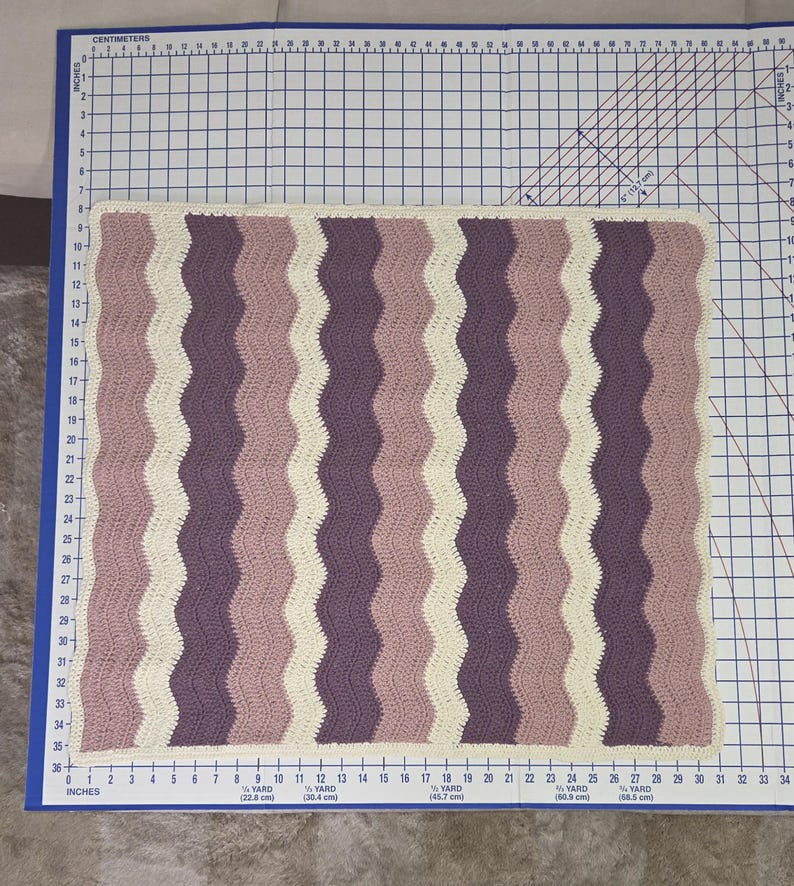 Pode incluir: Uma manta retangular de croch&ecirc; com um padr&atilde;o ondulado e listras verticais alternadas em creme, rosa antigo e roxo escuro. A manta est&aacute; posicionada sobre uma base de corte com medidas m&eacute;tricas, medindo aproximadamente 61 cm por 66 cm.