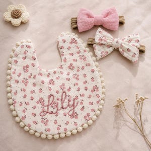 Puede incluir: Un babero para bebé y dos diademas con estampados florales rosas. El babero está bordado con el nombre "Lily" en hilo rosa y ribeteado con un ribete de pompón crema. Las diademas tienen lazos rosas.