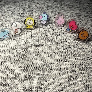 May include: A collection of seven small, clear acrylic pins featuring colorful cartoon characters. The set includes a light blue koala, a white dog, a yellow puppy, a light blue horse, a pink rabbit, a red heart, and a brown cookie character.