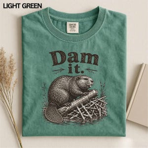 May include: A light green crew neck t-shirt featuring a dark brown graphic illustration of a beaver sitting on a log. The text "Dam it." is printed in a dark brown serif font above the beaver. The shirt has a Comfort Colors brand label.