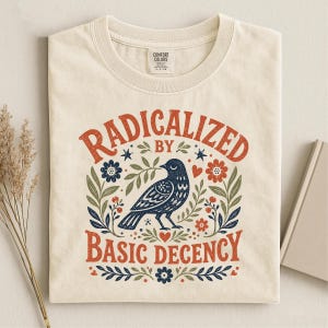 May include: A cream-colored t-shirt featuring a graphic print of a dark blue bird surrounded by green leaves, red flowers, and small stars. The text "RADICALIZED BY BASIC DECENCY" is printed in a curved, rustic orange font above and below the bird.