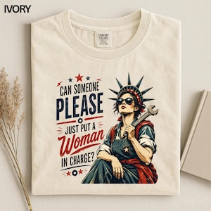 May include: An ivory crew neck t-shirt featuring a graphic of a person wearing a crown and sunglasses, holding a wrench. The text reads: CAN SOMEONE PLEASE JUST PUT A WOMAN IN CHARGE? The design uses red, white, and blue colors with star accents.