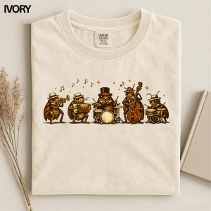 May include: An ivory-colored t-shirt featuring a graphic illustration of five anthropomorphic insects playing musical instruments, including a trumpet, accordion, drums, double bass, and percussion, accompanied by floating musical notes.