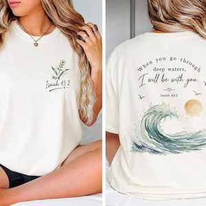 May include: A cream-colored t-shirt featuring a small floral graphic and the text Isaiah 43:2 on the front left chest. The back displays a large watercolor ocean wave, a sun, birds, and the text When you go through deep waters, I will be with you.
