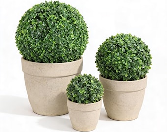 May include: Three artificial topiary plants in beige pots. The plants are spherical, with dense, vibrant green foliage. The pots are of varying sizes, creating a tiered effect. The background is plain white, highlighting the plants.