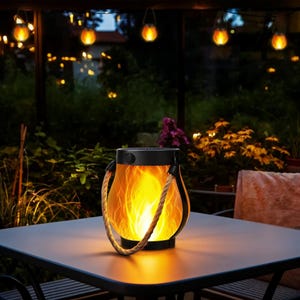 May include: A solar-powered lantern with a flame effect sits on a table. The lantern has a black top and base, with a clear body displaying an orange flame design. A rope handle arches over the top. String lights hang in the background.