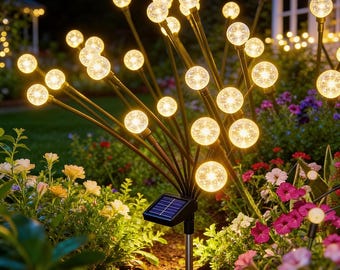 May include: Solar-powered garden lights with multiple glowing orbs on dark stems, resembling fireworks. The lights are set in a flower bed with various colorful flowers and greenery. A small solar panel is visible.